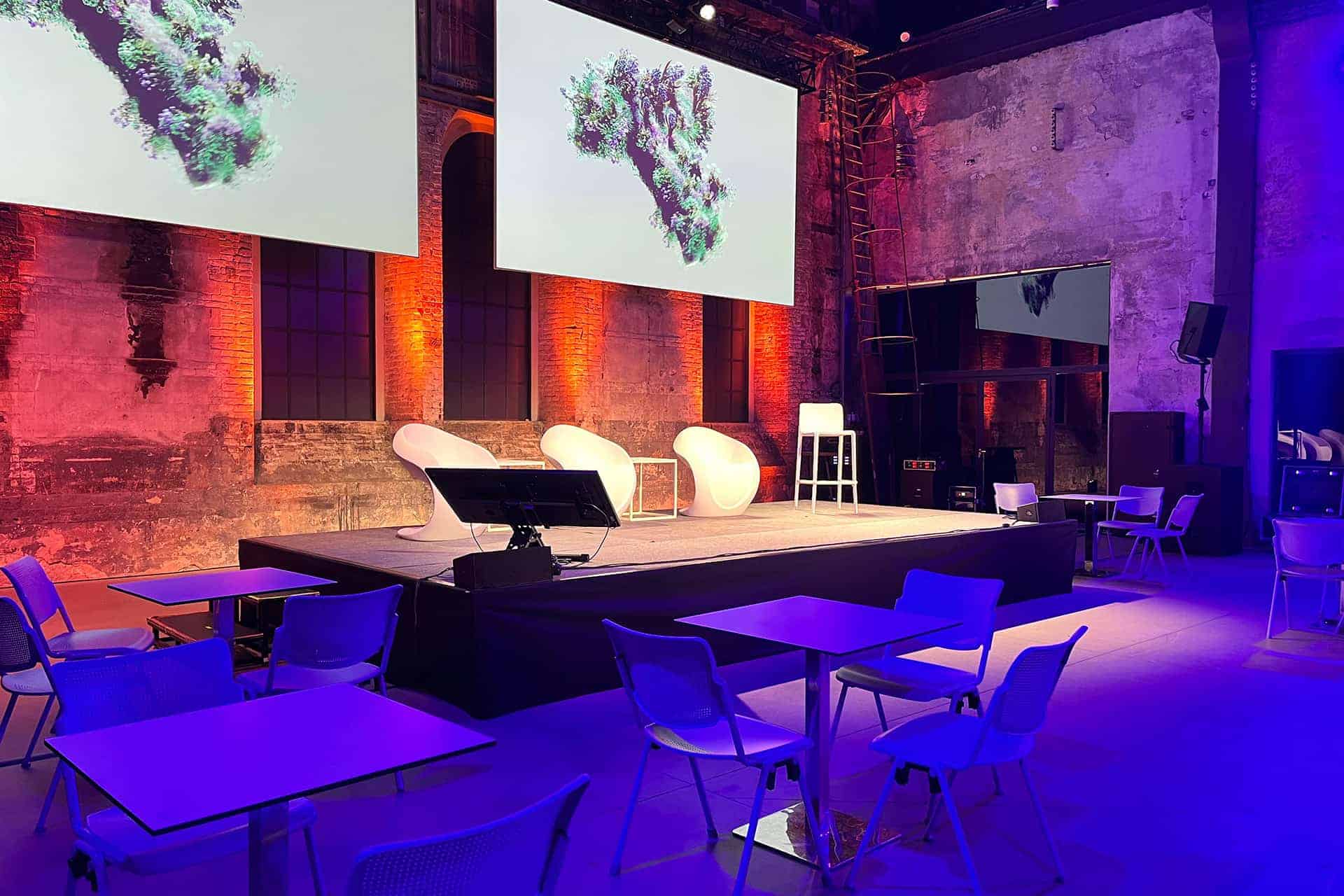 Palco per eventi con tre sedie moderne, due schermi in alto che trasmettono immagini astratte, circondato da tavoli e sedie in uno spazio scarsamente illuminato con pareti in mattoni a vista.