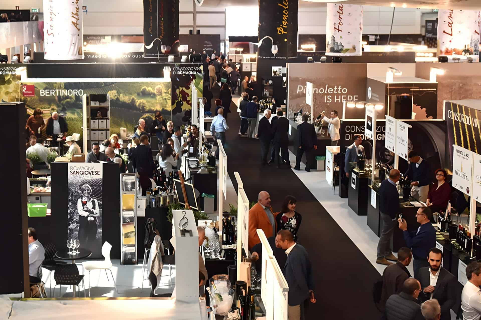 Vinitaly, la fiera dei professionisti del mondo del vino, con partecipanti che camminano tra gli stand che espongono vari marchi di vino. Gli espositori presentano striscioni e bottiglie, con persone che interagiscono e fanno networking nella sede ben illuminata.