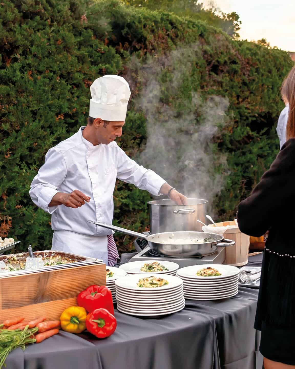 Chef professionista in abbigliamento bianco prepara piatti su un buffet all'aperto con verdure e stoviglie, in un ambiente naturale.