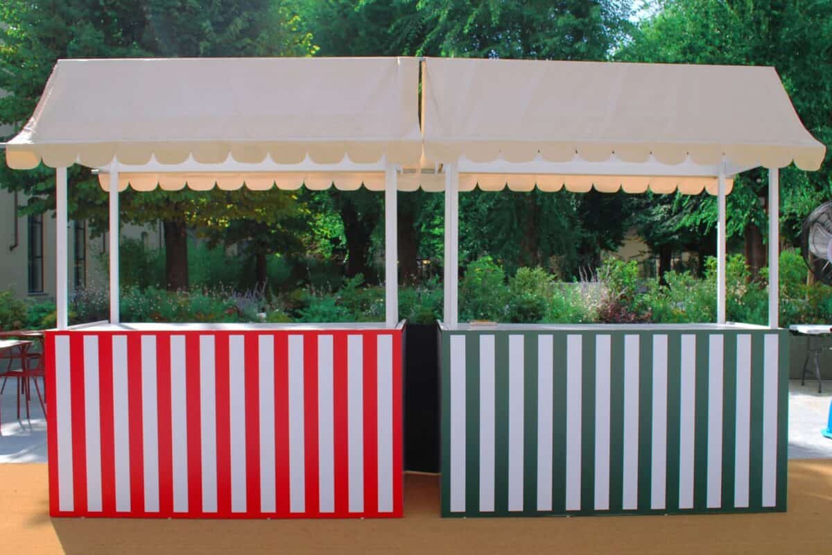 Stand gastronomico con tende bianche e pannelli a righe rosse e verdi, ideale per eventi all'aperto e fiere, con un ambiente naturale sullo sfondo.
