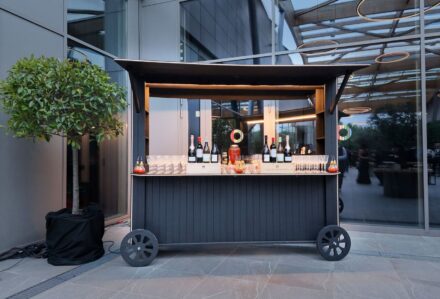 Baracchino mobile per eventi con bottiglie di vino e bicchieri, allestimento elegante in stile moderno, posizionato all'esterno di un edificio con vetrate e struttura in metallo.