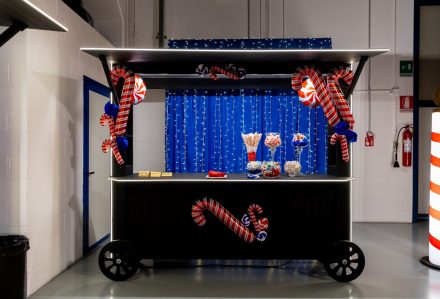 Lo stand "Bancarella Gourmet" è decorato con bastoncini di zucchero e luci, e presenta barattoli di dolciumi assortiti sullo sfondo di una tenda blu illuminata da lucine.