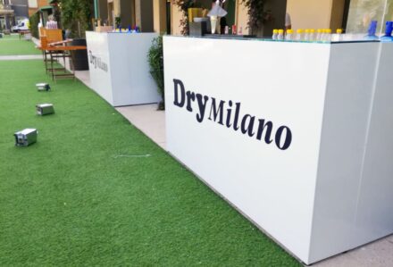 Pannello bar bianco con logo DryMilano, ideale per eventi e feste, posizionato su un prato artificiale verde, perfetto per creare un'area accogliente e moderna per il bancone White Russian.