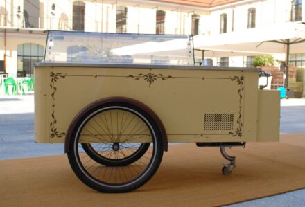 Carretto dei gelati vintage con tettoia a righe, struttura crema decorata e vetrina refrigerata, posizionato all’esterno.