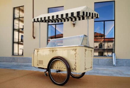 Carretto dei gelati vintage con tettoia a righe, struttura crema decorata e vetrina refrigerata, posizionato all’esterno.