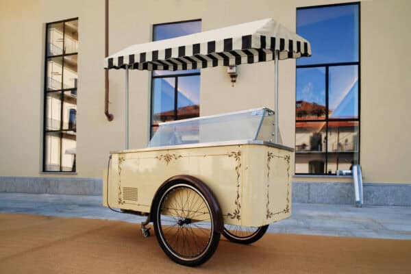 Carretto dei gelati vintage con tettoia a righe, struttura crema decorata e vetrina refrigerata, posizionato all’esterno.