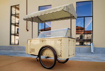 Carretto dei gelati vintage con tettoia a righe, struttura crema decorata e vetrina refrigerata, posizionato all’esterno.