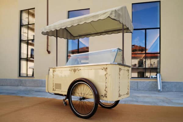 Carretto dei gelati vintage con tettoia a righe, struttura crema decorata e vetrina refrigerata, posizionato all’esterno.