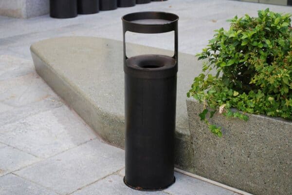 Cestino porta rifiuti nero in metallo con manico, posizionato su pavimentazione in pietra, vicino a una pianta verde in un'area pubblica.