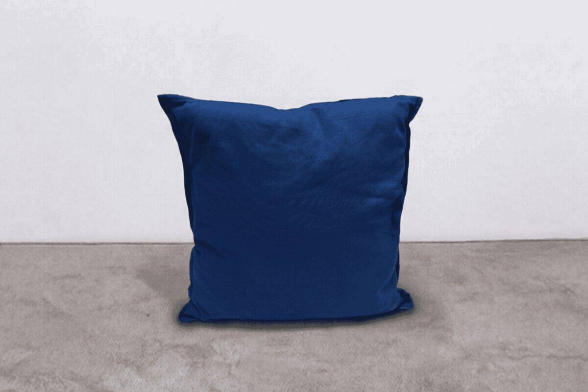 Cuscino blu in tessuto morbido posizionato su un pavimento in cemento, sfondo bianco semplice.