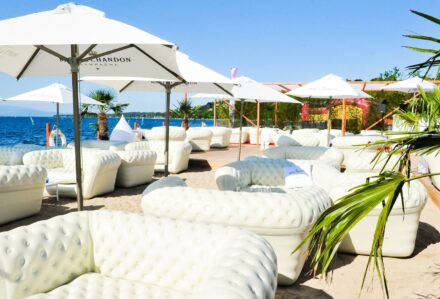 Divano gonfiabile grande bianco posizionato sulla spiaggia con ombrelloni e vista mare, ideale per relax e comfort all'aperto.