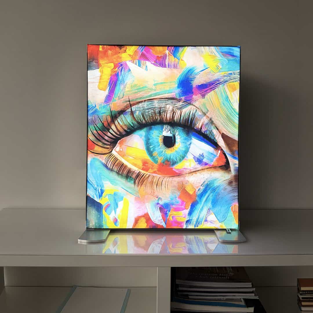 Quadro artistico con un grande occhio blu, dipinto con colori vivaci e astratti, posizionato su una superficie bianca.
