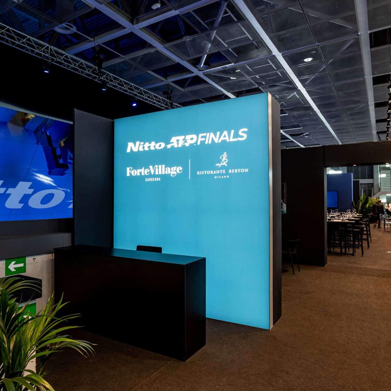 Stand di evento con pannello luminoso Nitto ATP Finals, Forte Village e Ristorante Burton, all'interno di una sala conferenze moderna.
