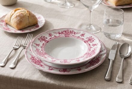 Set di stoviglie in porcellana con motivo floreale rosa e bianco, posate in acciaio inox, bicchieri e pane su tovaglia beige.
