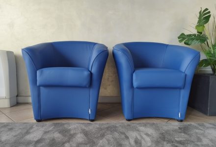 Due Poltrona Santorini in ecopelle blu sono posizionate una accanto all'altra su un tappeto grigio contro una parete neutra, con una pianta in vaso visibile sulla destra.