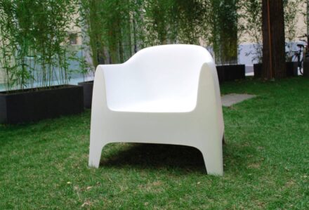 Sedia moderna bianca in plastica su prato verde con piante e alberi sullo sfondo.