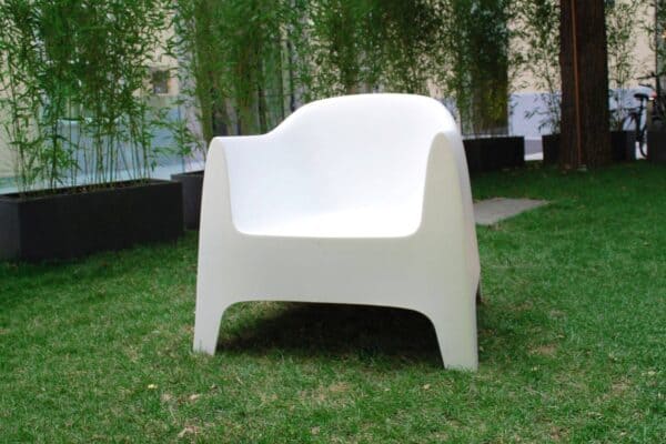 Sedia moderna bianca in plastica su prato verde con piante e alberi sullo sfondo.