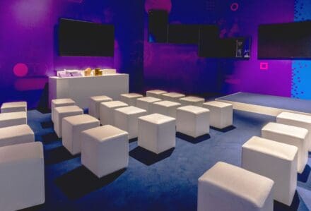 Sala conferenze moderna con sedie bianche e schermi grandi, illuminata con luci blu e viola, ideale per eventi aziendali e presentazioni.