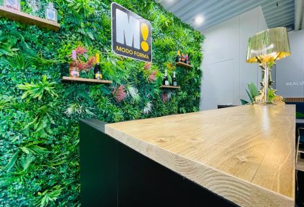 Un bar con un bancone in legno, una parete Quadrotta in Verde Artificiale da Parete, con mensole con bottiglie, e una lampada decorativa.