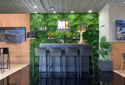 Uno stand espositivo con la Quadrotta in Verde Artificiale da Parete, un bancone nero con sgabelli e un arredamento moderno. Il logo "Modo Forma" è al centro.