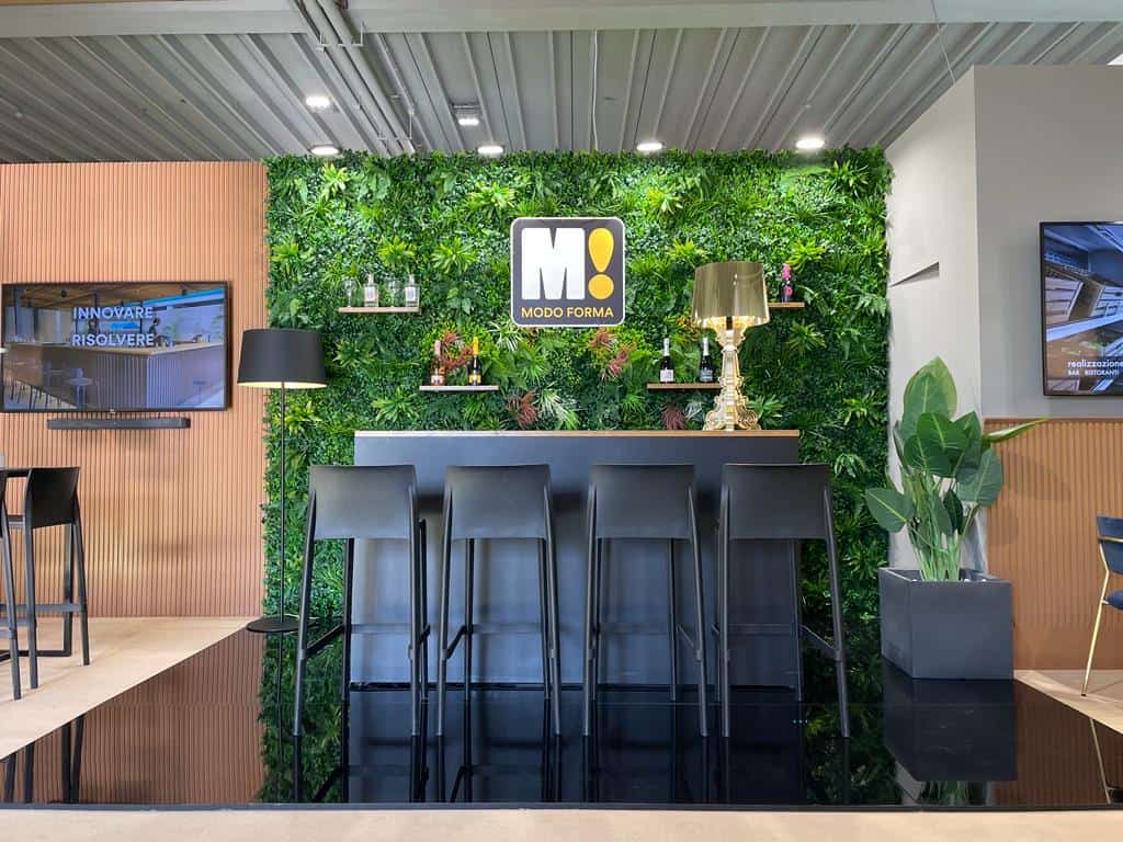 Uno stand espositivo con la Quadrotta in Verde Artificiale da Parete, un bancone nero con sgabelli e un arredamento moderno. Il logo "Modo Forma" è al centro.
