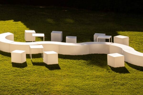 Seduta modulare bianca a forma di serpente con sgabelli abbinati su prato verde, ideale per eventi all'aperto e ambienti moderni.