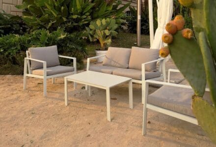 Set di mobili da esterno con divano e sedie in tessuto grigio e tavolino bianco in un giardino soleggiato.