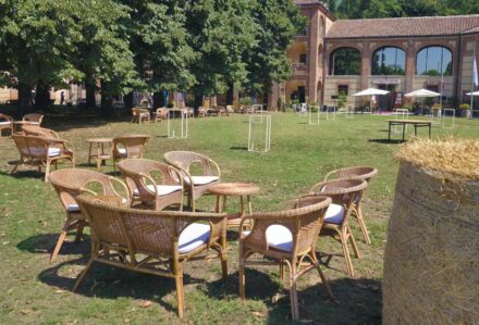 Sedie in vimini e tavoli all'aperto in un giardino con edificio storico sullo sfondo, ideale per eventi e feste all'aperto.