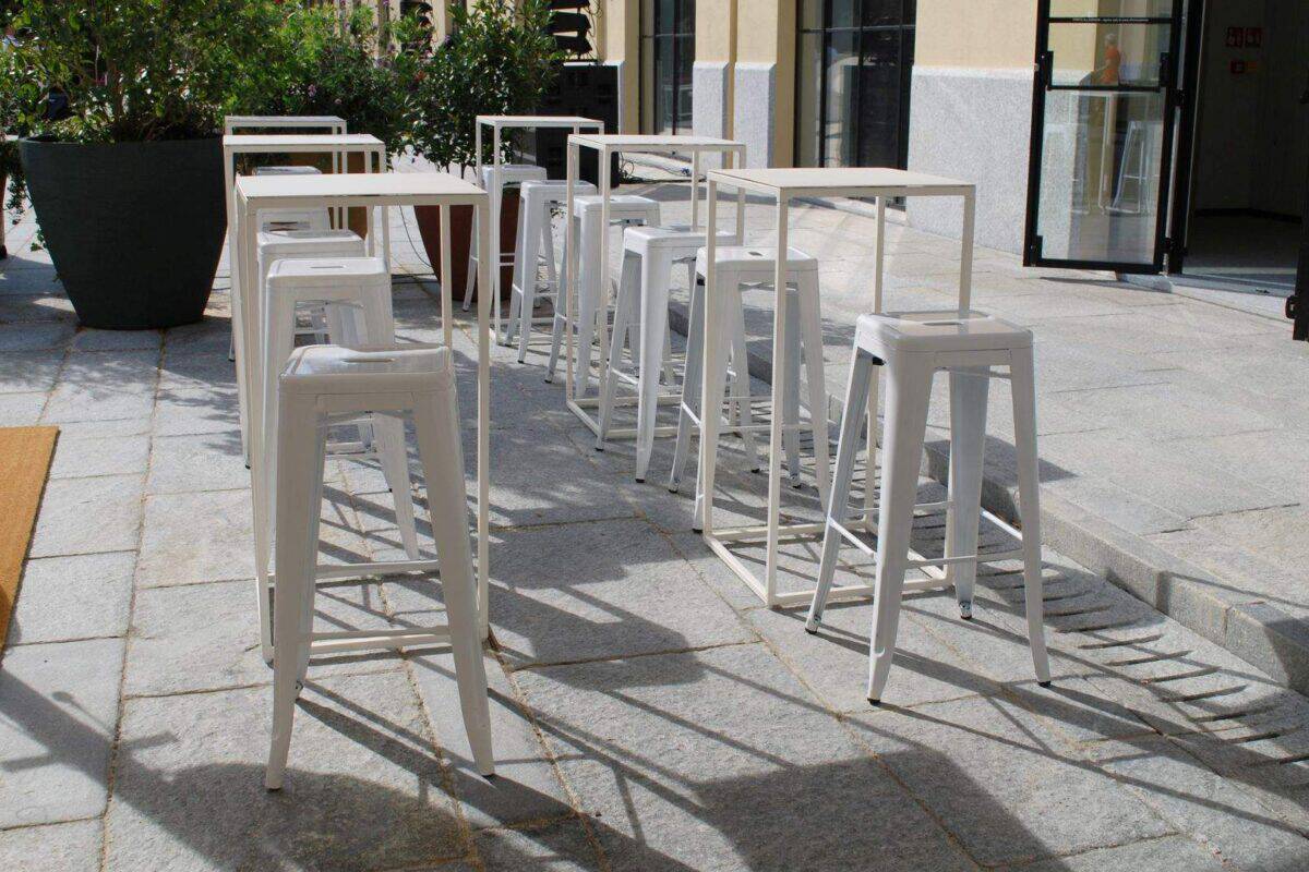 Sedie alte bianche in stile moderno disposte all'aperto su una terrazza in pietra, con piante e vetrate sullo sfondo, perfette per eventi o caffè all'aperto.