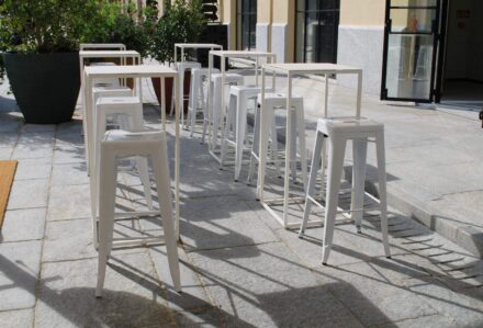 Sedie alte bianche in stile moderno disposte all'aperto su una terrazza in pietra, con piante e vetrate sullo sfondo, perfette per eventi o caffè all'aperto.