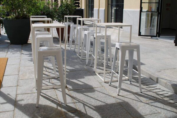 Sedie alte bianche in stile moderno disposte all'aperto su una terrazza in pietra, con piante e vetrate sullo sfondo, perfette per eventi o caffè all'aperto.
