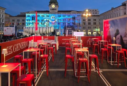 Area all'aperto con sedie rosse e tavoli in un evento serale in piazza, con edifici storici e proiezioni luminose sullo sfondo.