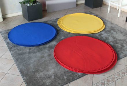 Tre pile di tappetini circolari in feltro blu, giallo e il "Tappeto rotondo diam. 100 cm rosso" sono posizionati su un tappeto grigio su un pavimento piastrellato.