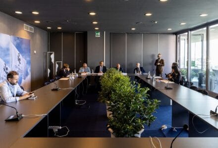 Sala riunioni moderna con partecipanti seduti a un tavolo lungo, piante al centro e grandi finestre che lasciano entrare luce naturale.
