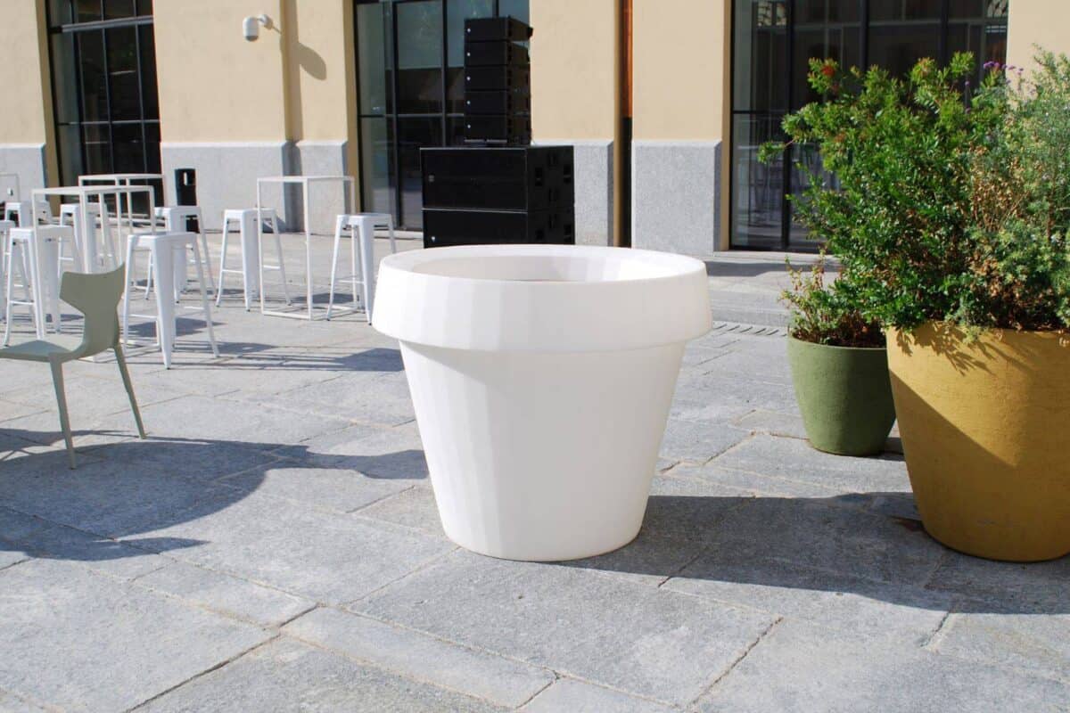 Vaso di grandi dimensioni in stile moderno, bianco, posizionato su una pavimentazione esterna con sedie e tavoli in background.