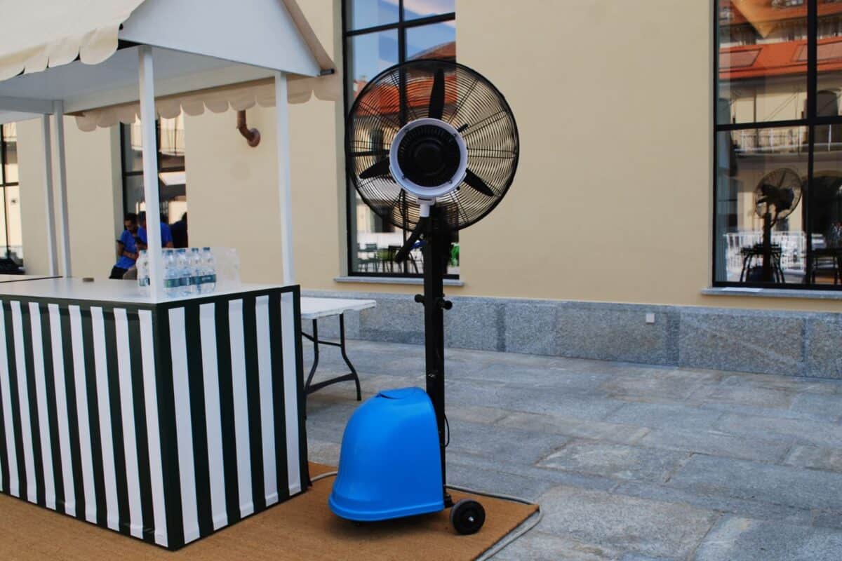 Ventilatore portatile con base blu e schermo nero, posizionato su pavimento in pietra in un'area esterna.