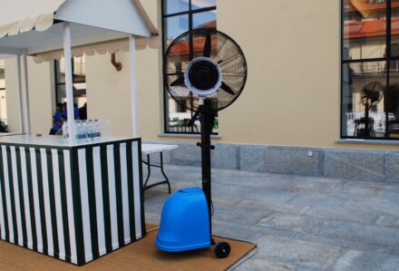 Ventilatore portatile con base blu e schermo nero, posizionato su pavimento in pietra in un'area esterna.