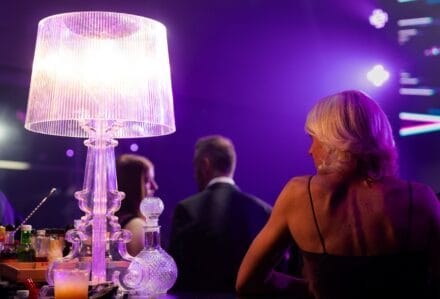 LAMPADA DA TAVOLO ELEGANTE IN VETRO TRASPARENTE CON LUCE CALDA, ATMOSFERA DI EVENTO SERIO E CHIC, CON PARTECIPANTI IN ABITI FORMALI IN UN'AMBIENTE CON ILLUMINAZIONE COLORATA.
