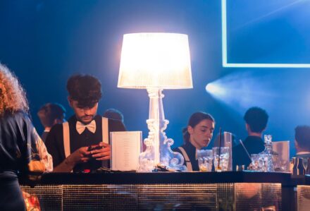 Elegante bar con clienti che sorseggiano drink, illuminato da lampade di design in un ambiente sofisticato e moderno, ideale per eventi e socializzazione di classe.