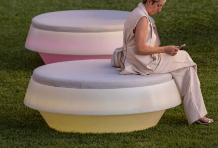Donna anziana seduta su una panchina moderna in un parco verde, che utilizza il suo smartphone, ambiente all'aperto con erba ben curata.