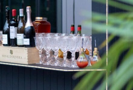 Schieramento di calici da vino modello Melodia in vetro intagliato su un bancone bar con bottiglie, disponibili a noleggio con Modorent per catering ed eventi.