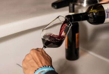 Persona versa vino rosso in un bicchiere con una bottiglia di vino rosso in primo piano, ambientato in una cucina moderna.